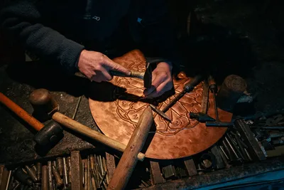 Un artesano elaborando meticulosamente un plato de metal detallado usando herramientas tradicionales en un taller con poca luz.